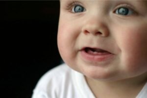 Bebê sorrindo com os primeiros dentinhos inferiores visíveis, usando uma camiseta branca e olhando com expressão curiosa.