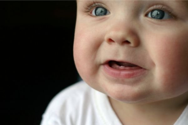 Bebê sorrindo com os primeiros dentinhos inferiores visíveis, usando uma camiseta branca e olhando com expressão curiosa.