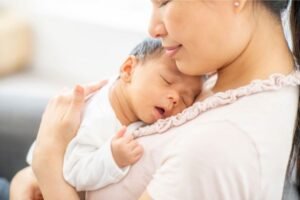 Mãe segurando seu bebê nos braços, com o pequeno dormindo tranquilamente encostado em seu peito, transmitindo carinho e proteção.