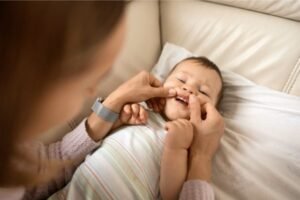 Mãe examinando a boca de seu bebê, que está deitado e sorrindo, enquanto ela levanta suavemente o lábio superior para verificar os primeiros dentinhos.