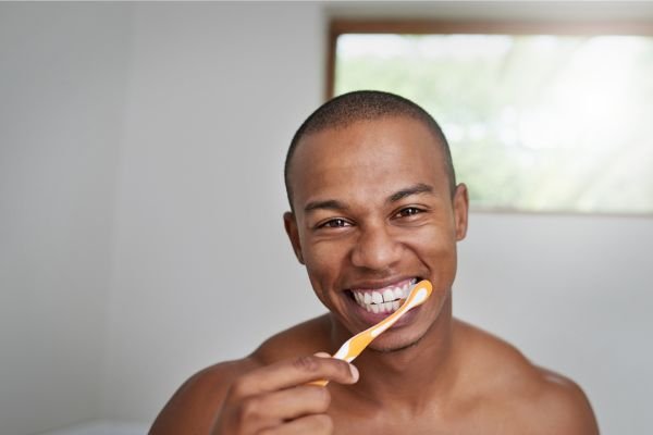 Homem sorrindo escovando os dentes com uma escova de cabo laranja em um ambiente iluminado, destacando bons hábitos de higiene bucal.