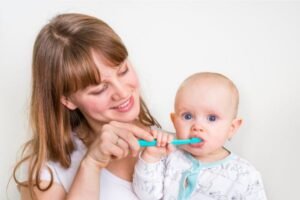 Mãe sorridente ajudando seu bebê a escovar os dentes com uma escova infantil azul, demonstrando cuidado com a higiene bucal desde cedo.