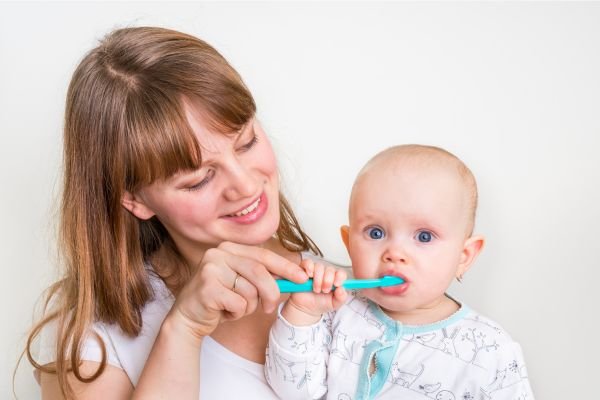 Mãe sorridente ajudando seu bebê a escovar os dentes com uma escova infantil azul, demonstrando cuidado com a higiene bucal desde cedo.
