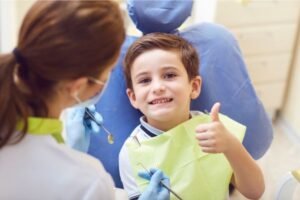 Criança sentada em uma cadeira odontológica, sorrindo e fazendo sinal de positivo com a mão, enquanto uma dentista com luvas e máscara segura instrumentos para exame.