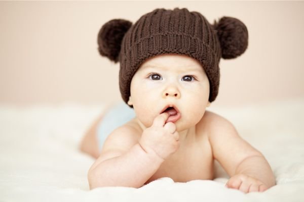 Bebê deitado de bruços em um cobertor macio, usando um gorro marrom com pompons, colocando o dedo na boca em um gesto curioso.