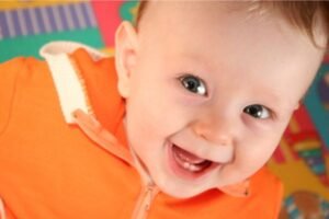 Bebê sorridente com os primeiros dentinhos visíveis, vestindo uma roupa laranja, em um ambiente colorido e alegre.