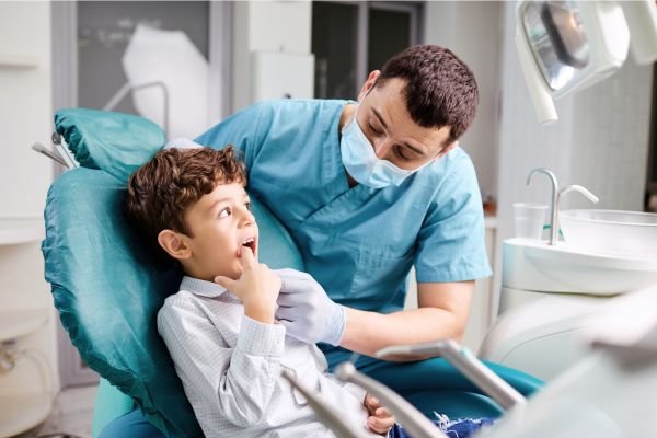Criança sentada em uma cadeira odontológica apontando para a boca, enquanto um dentista de máscara e luvas conversa com ela em um ambiente clínico.