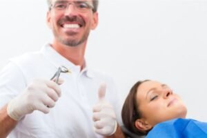 Dentista sorridente segurando um fórceps com um dente recém-extraído, fazendo sinal de positivo, enquanto um paciente relaxa na cadeira odontológica.