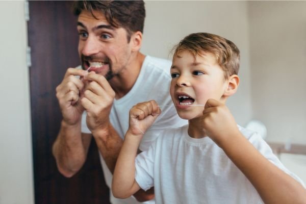 Pai e filho sorridentes usando fio dental em frente ao espelho do banheiro, ambos vestidos com camisetas brancas, demonstrando hábitos saudáveis ​​de higiene bucal.