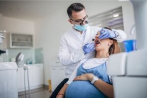 Mulher grávida sentada em uma cadeira odontológica sendo examinada por um dentista com máscara, luvas e jaleco branco, em um consultório moderno e bem iluminado.