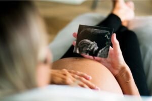 Mulher grávida deitada, segurando uma imagem de ultrassom e tocando a barriga, simbolizando expectativa e conexão com o bebê, enquanto destaca a importância de prevenir gengivite na gravidez.