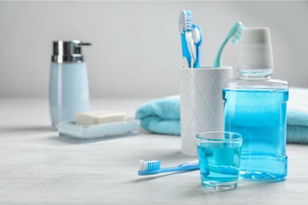 Conjunto de itens de higiene bucal em um banheiro, incluindo um enxaguante bucal azul, copo com escova de dentes, macarrão, toalha e líquido, organizados sobre uma bancada branca.