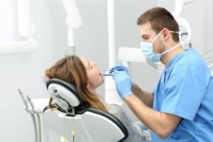 Dentista realizando um exame em uma paciente sentada na cadeira odontológica, usando máscara, luvas e instrumentos para inspeção bucal.