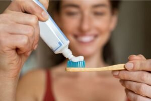 Mulher sorridente aplicando creme dental em uma escova de dentes de cabo de madeira, destacando hábitos saudáveis de higiene bucal.