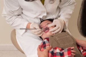 Bebê deitado no colo de um responsável durante uma consulta odontológica. O dentista, usa jaleco branco e luvas, examina a boca do bebê com um espelho dental.