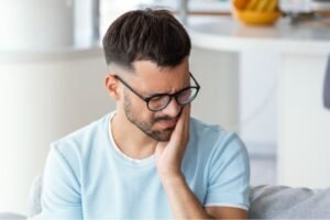 Homem com expressão de dor segurando o lado do rosto, sugerindo desconforto ou dor de dente, sentado em um ambiente doméstico.