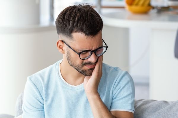 Homem com expressão de dor segurando o lado do rosto, sugerindo desconforto ou dor de dente, sentado em um ambiente doméstico.