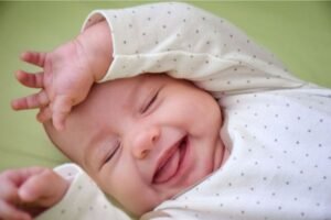 Bebê sorrindo de olhos fechados, com expressão feliz, usa roupa clara com estampas de bolinhas e deitado em um fundo verde claro.