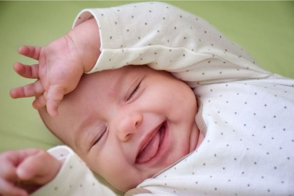 Bebê sorrindo de olhos fechados, com expressão feliz, usa roupa clara com estampas de bolinhas e deitado em um fundo verde claro.