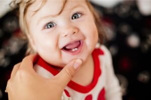 Bebê sorridente mostrando os primeiros dentinhos inferiores, vestindo roupa branca com detalhes vermelhos, enquanto uma mão segura suavemente seu queixo.